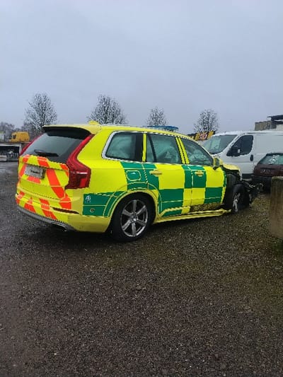 An exterior photograph depicts a heavily damaged yellow and green emergency vehicle, identified as a Volvo XC90 SUV, stationary on a dark gravel surface in Bitburg, Germany, under an overcast sky. The vehicle displays bright yellow and green Battenburg markings along its sides and rear quarter panels, with red and yellow diagonal chevron reflective tape across the lower portion of its tailgate. The front passenger side exhibits severe impact damage, with the bumper and fender assembly deformed or missing, and the front passenger wheel appears displaced and angled inwards. Minor scuff marks are present on the passenger-side door. In the background, a white cargo van and a partially obscured darker vehicle are visible. To the far left, a yellow industrial vehicle and a building structure can be seen, with several bare deciduous trees in the mid-ground. The scene is devoid of people or discernible activity.