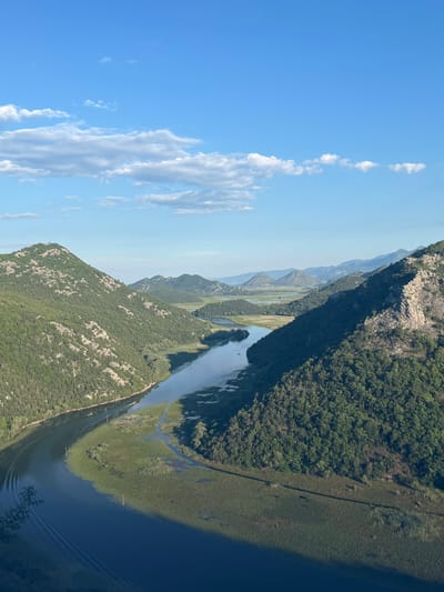 A high-angle, eye-level shot captures a serene, expansive view of a river winding through verdant, tree-covered hills under a bright blue sky with scattered white clouds. The river, a deep blue, meanders through the lush green landscape, with a boat visible on the water, leaving a faint wake. The surrounding hills are densely forested, with rocky outcrops interspersed throughout. In the distance, more rolling hills fade into a hazy, lighter green. The scene is devoid of any human presence besides the distant boat, creating a peaceful, natural panorama. The location is Rvaši, Montenegro.