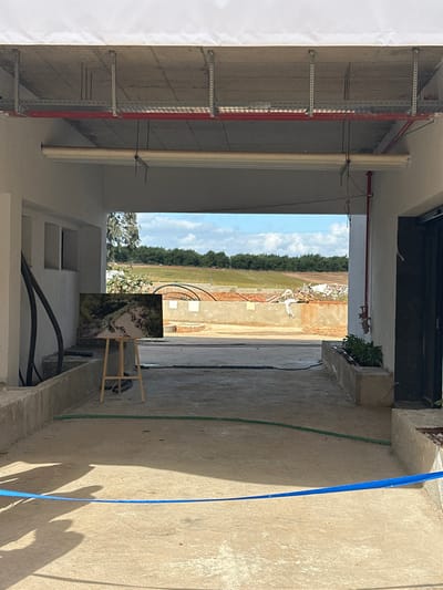 The scene depicts an interior view of an open-sided, partially constructed or industrial-style building situated in Kadima-Zoran, Israel, looking outwards towards an open, undeveloped landscape under a bright, partly cloudy sky.
The immediate foreground features a light-colored concrete floor illuminated by sunlight, casting prominent shadows from unseen entities on the lower left. A blue fabric ribbon or tape runs horizontally across the floor in the lower frame. On the left, a wooden easel displays a horizontal artwork or photograph depicting a rocky stream flowing through a vegetated area. Behind the easel, multiple black flexible pipes are arranged against a light-colored wall containing three rectangular recessed openings. The ceiling is exposed concrete, revealing overhead infrastructure including multiple parallel metal cable trays and a red fire suppression pipe running horizontally. A white roller blind mechanism is also mounted below the ceiling structures. On the right, a vertical red pipe is affixed to a light-colored wall, extending downwards towards a small concrete planter box containing green vegetation. A dark door frame is partially visible on the far right. A green garden hose is coiled on the concrete floor, extending towards the outdoor area.
The midground, visible through the building's opening, consists of a flat, unpaved area that appears to be a construction site or undeveloped land. Low concrete barriers or structures are present, along with scattered debris including pieces of wood and white sheeting, particularly on the right. Large, curved black flexible pipes are noticeable on the left side of this outdoor expanse.
The background reveals a panoramic view of open fields, characterized by areas of brown earth and green vegetation, indicating agricultural or natural terrain. A dense line of dark green trees forms the distant horizon, beneath a bright blue sky dotted with scattered white cumulus clouds.
