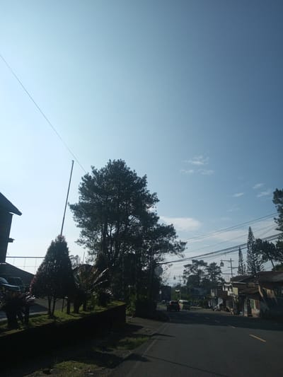 The image shows a street scene with trees, buildings, and power lines against a blue sky with some clouds. The road curves slightly to the right. There's a vehicle in the distance on the road. On the left side of the road, there are trees and a low wall. Buildings are visible on the right side of the road. There is no way to know the location is Phouviang-Gnai, Laos, or anywhere in Laos. There is no information about people.