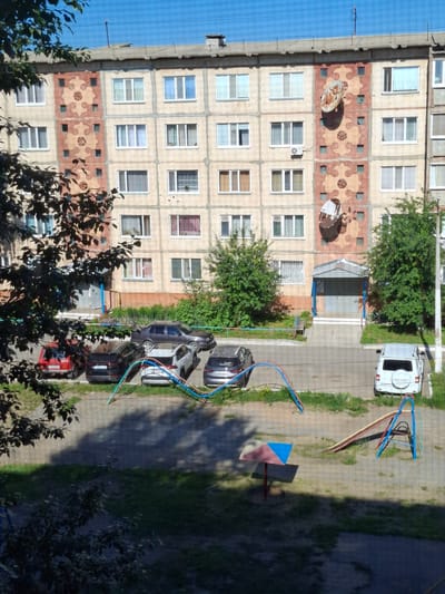 This image depicts an outdoor scene in the city of Jitiqara, Kazakhstan. In the foreground, there is a playground area with a colorful umbrella-like shade structure and a slide with a metal frame. Grass and patches of dirt surround the playground equipment.
Dominating the middle ground is a parking lot with several cars parked. Visible vehicles include a red sedan, a black SUV, a white SUV with the word "PATRIOT" on the back, and a dark-colored sedan. Some of these cars are partially obscured by foliage. Colorful playground safety barriers, consisting of blue, green, and red poles with wires, are present between the parking area and the playground.
The background features a multi-story apartment building. The building has a light-colored facade with decorative elements, including geometric patterns and circular motifs in shades of pink and beige. Several windows with white frames are visible on the building's exterior. Two satellite dishes are attached to the building's facade, positioned vertically. An awning with blue supports covers the entrance to the building. Trees with green foliage are situated to the left and right of the building entrance, and also along the building's facade. The sky above is clear and blue.