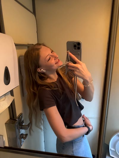 A young woman with long blonde hair takes a selfie in a small, well-lit room with cream-colored walls. She is wearing a dark, cropped t-shirt and light-wash denim jeans. She has white earbuds in her ears and is holding a grey smartphone with a multi-camera system in her right hand. A grey lanyard is attached to the phone case. She is wearing a beaded bracelet on her left wrist and a black smartwatch on her right wrist. A white paper towel dispenser is visible on the left side of the image. A metal fixture, possibly a coin slot or a lock, is mounted on the wall to the left of the woman. A mirror reflects the woman and the room, revealing a toilet on the far right. The environment suggests a public restroom or a small, utilitarian space. The location of Nizhnyaya Baskaya, Russia, is not discernible from the image.