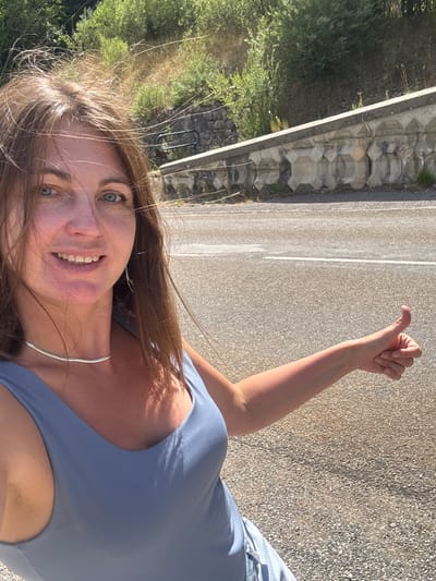 The media file is a selfie taken outdoors in a vertical orientation. A young woman with long brown hair and blue eyes is the primary subject. She is smiling and looking at the camera. She is wearing a light blue tank top and a silver necklace. Her right arm is raised with her thumb up, suggesting she is hitchhiking or signaling.
The setting appears to be a roadside, with a paved road and a gravel shoulder visible. To the left of the road, there is a stone wall and lush green vegetation, including trees and bushes. The sun is casting shadows, indicating it is daytime, likely late afternoon or early morning due to the warm lighting. The overall mood is cheerful and adventurous.
There is no visible text in the image. The location context of Hauts de Bienne, France, suggests a rural or mountainous area, which is consistent with the natural scenery depicted. The activity taking place is the woman taking a selfie while likely on a journey.