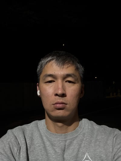 A close-up, waist-up, front-facing selfie shows a person with short, graying hair and light skin. The person is wearing a light gray crewneck sweatshirt. A white wireless earbud is visible in the left ear. The background is dark and appears to be outdoors at night, with a faint light visible in the distance. The scene is a city, but the specific location in Vancouver, United States of America cannot be determined from the image.