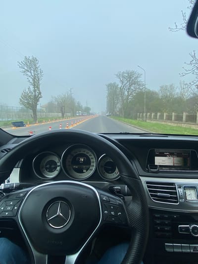 The image depicts the interior of a Mercedes-Benz car, focusing on the dashboard and steering wheel. The speedometer reads 74 km/h, with the car navigating a road marked by orange and white traffic cones, possibly indicating a construction zone. The infotainment system displays FM radio station "BNR BG RADIO" at 96.9 MHz. Through the windshield, a foggy, overcast day is visible, with trees lining the road and power lines overhead. Based on the limited information and the OCR text, the location could be Vetrino, Bulgaria, indicated by the radio station "BNR" (Bulgarian National Radio).