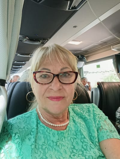 An elderly Caucasian woman with blonde hair and glasses is taking a selfie on a bus. She is wearing a teal lace top and a pearl necklace. The bus has black seats and a curtain on the left side. In the background, other passengers can be seen. The lighting suggests it is daytime. The image appears to be taken in Terras de Bouro, Portugal, as indicated by metadata.