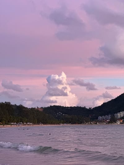 The image depicts a coastal scene with a mountain covered in lush green trees and buildings on the left side, and a calm ocean with gentle waves on the right. The sky is a gradient of soft pinks and purples, suggesting either sunrise or sunset. Scattered clouds add texture to the sky.
Along the coastline, a beach is visible, lined with palm trees and a few scattered buildings. There are small groups of people on the beach and in the water, appearing to be enjoying a leisurely time. One person is visible in the water, possibly swimming or playing. A dark, round object, potentially a ball, is floating in the water.
The overall mood is serene and peaceful, bathed in the soft hues of twilight. The location is identified as Ban Bang Thao, Thailand, a popular beach destination. No visible text is present in the image.