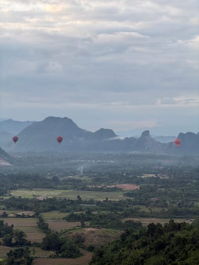 This is a vertical, medium-distance shot of a misty, mountainous landscape, likely at sunrise or sunset in Ban Phon Sai, Laos. Four red hot air balloons are visible against a backdrop of lush green hills and valleys. The balloons are at varying altitudes, with three closer to the left and center of the frame and one further to the right. The foreground is dominated by dense green foliage, with agricultural fields and some scattered buildings visible in the midground. The background features layers of mist-shrouded mountains. The sky is overcast with pale, diffused light. There are no people clearly visible in the image.