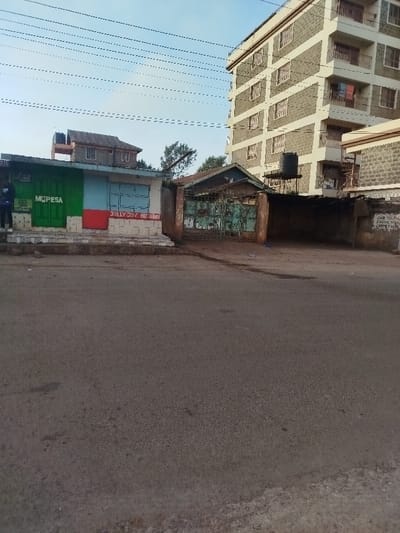A street view features a low building with "M-PESA" and "JOLLY JOY BUTCHERY" signs. An adjacent gate with an overgrown entrance sits next to a weathered wall with illegible writing. A multi-story building looms behind. Power lines crisscross the sky, and the road occupies a significant portion of the foreground. A smaller building with a water tank on its roof is behind the lower structure.
