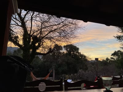 The image captures a sunset view from a covered outdoor area in El Rodeo, Argentina. In the foreground, a dark, amorphous object, possibly a bag or backpack, rests on a wooden surface. Two dark wooden chairs with circular cutouts are visible, facing outwards towards the scenery. To the right, a clear glass with a dark rim sits on the wooden table. Beyond the chairs, a hammock with an orange net is suspended between two posts. A tall, leafless tree with ivy climbing its trunk dominates the left side of the frame, its branches silhouetted against a sky painted with hues of orange, yellow, and blue from the setting sun. The background features a dense forest and distant hills. A slender metal pole with a square fixture on top stands in the middle ground. The overall scene depicts a tranquil outdoor setting at dusk.