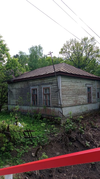 The image depicts an aged, single-story wooden house located in Дубки, Russia. The exterior paint of the house is peeling, revealing bare wood underneath. The roof is made of metal sheets, and an old, weathered telephone pole leans against the side of the house. Two windows with red frames are visible on one side of the house, while another window is located on the adjacent side. The surrounding area is overgrown with vegetation, including trees, shrubs, and various plants. A red metal railing is visible in the foreground. The sky is overcast, providing diffused lighting.
