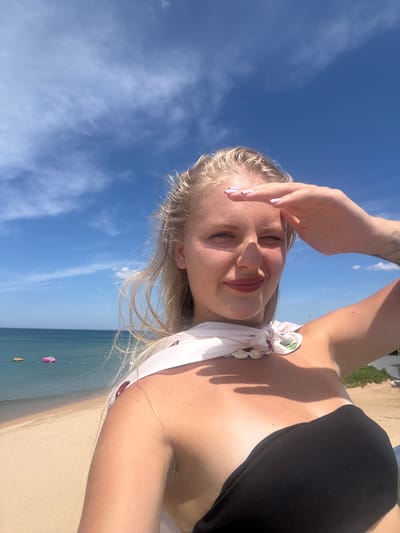 A young woman with blonde hair and fair skin is taking a selfie on a sunny day at the beach. She is wearing a black strapless top and a white scarf adorned with seashells around her neck. Her right hand is raised to her forehead, shielding her eyes from the sun as she looks towards the camera. The background shows a sandy beach, the turquoise ocean, and a clear blue sky with scattered clouds. In the distance, two inflatable objects, possibly water toys, are visible on the water. The scene appears to be in Tuy Hoa, Vietnam, a coastal city known for its beautiful beaches.