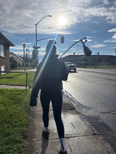 A person walks along a sidewalk on a sunny day in Thunder Bay, Canada. The person is viewed from behind, wearing dark athletic leggings and a dark hooded jacket. They are carrying a long stick with what appears to be a bag attached to the end. The sidewalk is cracked and adjacent to a grassy area on the left. Across the street, there is a road with a white pickup truck visible. The sky is blue with scattered white clouds, and the sun is shining brightly, creating lens flare. Streetlights and traffic signals are present. In the background, buildings and trees are visible. The road appears wet, possibly from recent rain, with reflections of the sky.