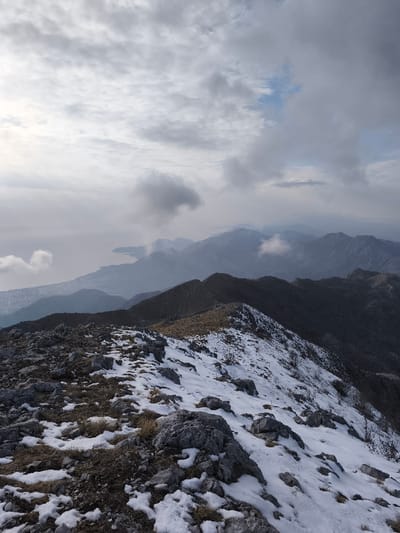 An elevated landscape photograph, taken from a rocky, snow-dusted mountain ridge in Mali Mikulići, Montenegro, depicts a panoramic view under an overcast sky. In the foreground, patches of snow intersperse with gray rocks and sparse, dry, brown vegetation along a rugged, undulating ridge line. A subtle path-like indentation is visible traversing the ridge. The midground features multiple successive mountain ranges, gradually fading into a blue-gray haze. To the far left in the background, a large body of water, likely the Adriatic Sea, stretches along a visible coastline, with a dense urban settlement situated along its shore. The sky is largely covered by varying densities of gray and white stratocumulus clouds, with some darker, thicker formations and occasional glimpses of lighter sky. No individuals or man-made structures other than the distant urban area are visible. The scene suggests a cold, high-altitude environment, possibly late autumn or winter.