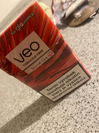 The image shows a close-up of a red and orange striped cigarette pack, labeled "Veo Tobacco Free" and "for glo HYPER". Below this, there is text in a light brown box that reads "Tento výrobek obsahuje nikotin, který je vysoce návykovou látkou." This translates to "This product contains nicotine, which is a highly addictive substance." The pack is placed on a speckled grey surface that resembles a countertop or table. The background is blurred but appears to be indoors. There are no visible people, animals, or specific landmarks that would place this image in the city of Jihlava, Czechia. The lighting suggests an indoor setting, possibly a home or retail environment.