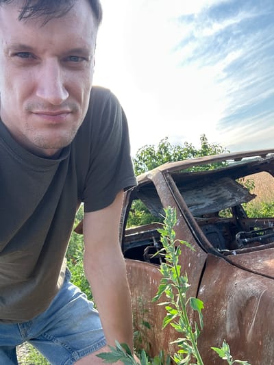 In Slobidka-Rakhnivska, Ukraine, a man in a dark green t-shirt and blue denim shorts is shown in a selfie. He is leaning forward, with his face close to the camera, looking directly at it. Behind him, the rusted hulk of a burnt-out car is visible, with its interior and roof heavily damaged. Green foliage and tall grass surround the wreckage, and a bright, partly cloudy sky is overhead. The man's expression is serious. The overall scene suggests a post-conflict environment.