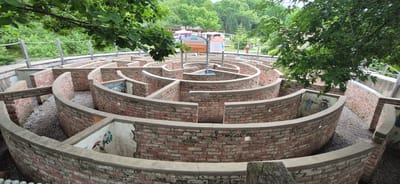 In Knevichi, Russia, an aerial view captures a circular brick maze with a concrete cap on its walls, situated within a lush, tree-lined park. The maze is designed with concentric pathways and intersecting walls, creating a complex network of routes. Several sections of the maze walls are adorned with faded murals depicting cartoon-like figures, including what appear to be gnomes and other fantastical characters. The ground within the maze is covered in small, gray pebbles. In the background, a wooden walkway with a rope railing winds through the greenery, suggesting it is an elevated path for observation or access to the maze. Above the maze, a sign with Cyrillic text is visible, indicating it is a tourist attraction. The overall scene depicts an outdoor recreational area, likely an amusement park or a dedicated maze attraction, designed for visitors to navigate and explore.