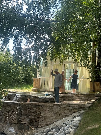 The image depicts three people standing outdoors in what appears to be a garden or a courtyard next to a modern building with large windows. The scene is oriented vertically, with the sky and treetops at the bottom and the ground at the top.

The subjects are two women and one man. The woman on the right is wearing a dark blue polka-dot dress and is holding hands with the woman in the middle, who is wearing a patterned dress. The man on the left is wearing a light-colored, checkered shirt and dark pants, and a light-colored hat. He is looking down and appears to be bowing or gesturing. The people seem to be engaged in some sort of interaction, possibly dancing or a ceremonial gesture, given their hand-holding and the man's pose.

The setting includes a building with a glass facade, a stone-paved pathway or terrace, and lush green trees and foliage. There is also a rock wall with water flowing down it, creating a small waterfall, and a grassy area to the right. The presence of flowing water suggests a possibly warm and sunny day, and the abundance of green suggests it is likely spring or summer. The lighting indicates it is daytime, with sunlight filtering through the leaves.

There is no visible text in the image. The overall mood seems pleasant and serene, with the natural elements of trees and water contributing to a tranquil atmosphere. The architecture of the building suggests a modern or perhaps a residential setting. The location context provided is Bogdanikha, Russia.