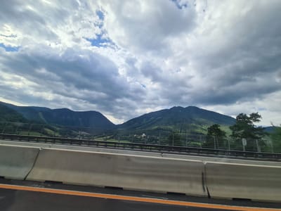 This image captures a view from a highway in Breitenstein, Austria, looking towards rolling green mountains under a cloudy sky. In the foreground, a concrete highway barrier with an orange and black striped lane marker is visible. Beyond the barrier, a metal fence separates the highway from a grassy slope with scattered trees. Further in the distance, lush green mountains dominate the landscape, with some areas appearing as clear-cut slopes and others densely forested. A small building, possibly a castle or a large house, is nestled in the valley between the mountains. The sky is filled with dramatic, heavy clouds, with patches of blue sky peeking through, suggesting dynamic weather conditions. The overall scene conveys a sense of a scenic drive through the Austrian countryside.