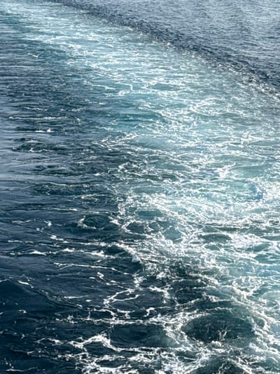 The image captures a detailed view of open water off the coast of Kamariotissa, Greece. The scene is dominated by a prominent wake of foamy white and light blue water, indicating the recent passage or ongoing movement of a vessel, which itself is not visible. This turbulent trail extends diagonally from the lower right to the upper left of the frame. The water outside the wake is a darker, calm blue with subtle ripples. No people, vessels, or land features are present within the observable marine environment.