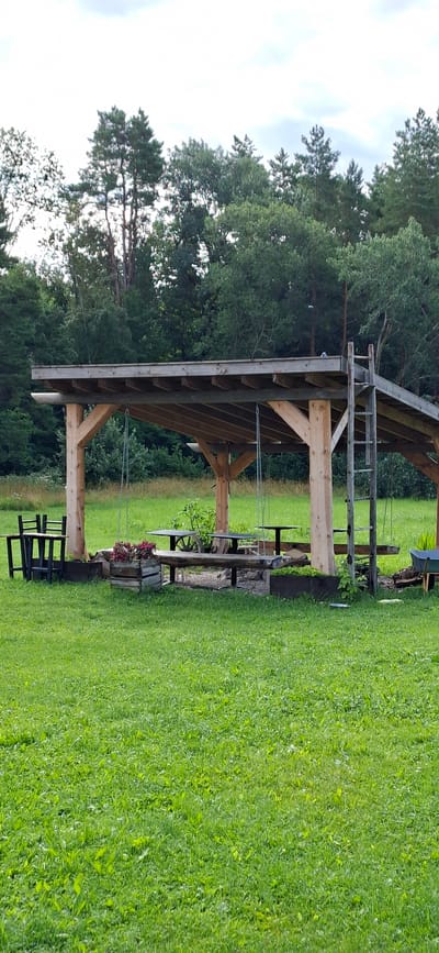 The image depicts an outdoor recreational area with a wooden gazebo. The gazebo has a flat roof supported by thick wooden posts. Inside, there are wooden benches and tables, and hanging swings. To the left of the gazebo, there are black wooden chairs and a table. A wooden crate filled with red flowers sits in front of the gazebo. In the background, a line of trees forms a dense forest. The ground is covered in lush green grass. The scene is likely located in a rural or park setting outside of a city. No people are visible in the image. The location is identified as Orissaare, Estonia.