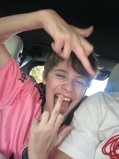 A young person inside a car gives the middle finger with both hands. They wear a pink soccer jersey with black stripes on the shoulders and appear to have a seatbelt on. They are sticking their tongue out and grimacing, with light shining through a car window in the background. Part of another person wearing a white shirt is visible at the top right of the frame.