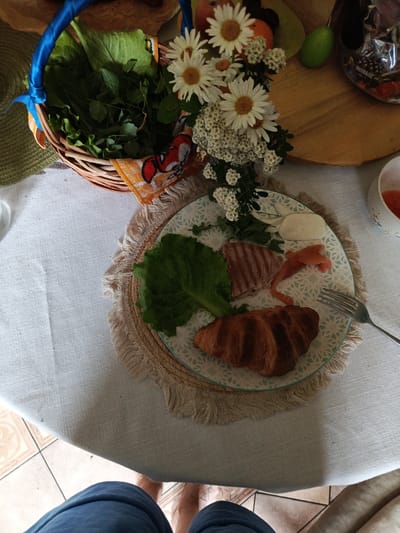 A high-angle shot shows a close-up of a breakfast spread laid out on a white tablecloth, with portions of a person's legs visible at the bottom. In the center of the frame, a round plate with a blue and white patterned rim sits on a fringed placemat. The plate holds a croissant, a slice of what appears to be cooked meat, a piece of smoked salmon, a dollop of white cheese, and a green lettuce leaf. A fork rests on the right side of the plate. To the left of the plate, a woven basket is filled with fresh green leaves and mint, adorned with a blue ribbon and a decorative orange and red patterned cloth. A bouquet of white daisies and small white flowers is positioned behind the basket. Further to the right, a wooden board holds a green egg-shaped object, some fruit (likely plums and an orange), and a clear plastic bag filled with chocolates. A white cup with a pink interior is partially visible on the far right. The background suggests an indoor setting with tiled flooring visible at the bottom. The scene is devoid of any specific identifiers placing it in Alexandrovskaya, Russia.