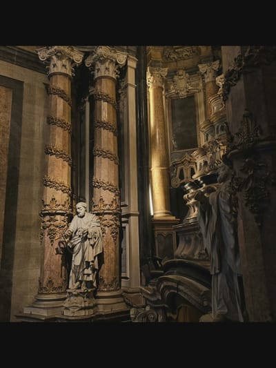 The image depicts the interior of a church, likely a basilica, with ornate Baroque architecture. Two marble statues of robed male figures are prominently featured. The statue on the left, positioned in the foreground, is of a bearded man holding a key and a chain, standing on a pedestal inscribed with Latin text. This is identified as Saint Peter. The statue on the right, further back and partially obscured, also appears to be of a similar religious figure, with hands clasped in prayer.

The statues are situated between massive, fluted columns adorned with gilded floral motifs and intricate capitals. The columns appear to be made of marble with reddish and beige veining. Behind the statues, the church's interior reveals more elaborate architectural elements, including gilded sculptures, niches, and arched structures. The lighting is dim, with a strong directional light source illuminating the right side of the scene, casting shadows and highlighting the textures of the marble and gold ornamentation.

The environment suggests a place of worship, with its grandeur and religious iconography. The location is identified as the Basilica di San Benedetto in Norcia, Italy.