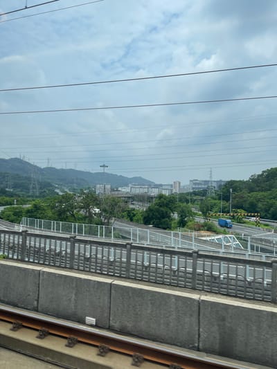 This media file captures a scene from a high vantage point, likely from a train or elevated walkway, looking out over a city and surrounding natural landscape. The perspective is from the side, with the image rotated 90 degrees counter-clockwise.

The primary subjects include a busy road with a blue vehicle, and a pedestrian walkway or bridge with a metal railing. Beyond this, the scene transitions into a lush, green hilly landscape dotted with buildings and transmission towers. The sky is overcast with clouds.

The image appears to be taken during the daytime, as evidenced by the lighting. The weather seems to be cloudy or possibly just after rain, contributing to a somewhat muted color palette. The presence of numerous power lines crisscrossing the frame suggests an urban or semi-urban environment with significant electrical infrastructure. The buildings in the distance indicate a developed area, while the hills suggest a location on the outskirts of a city or a region with varied topography.

No people are clearly visible. Notable details include the distinct structure of the transmission towers and the segmented concrete construction of the elevated walkway. The presence of buildings with "TCL" branding on them indicates a potential industrial or commercial area.

The overall emotion conveyed is one of observation and a sense of distance, looking out at the interplay between human development and the natural environment. The image provides a glimpse into the urban sprawl and infrastructure of a location like Guangming District, China.