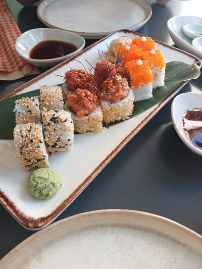 A triangular plate features assorted sushi. Varieties include rolls topped with orange tobiko roe, spicy tuna, and a mix of sesame seeds. Slices of ginger and decorative red threads garnish the platter. A smear of wasabi accompanies the sushi. Small bowls holding soy sauce and other plates frame the main dish, set on a gray surface. A red and white checked napkin sits nearby.