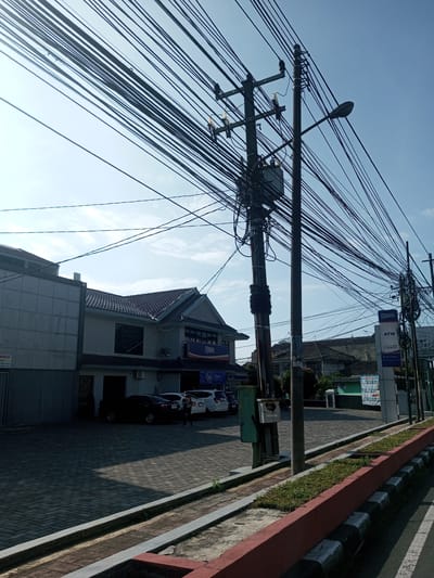 The image shows a street scene in an urban area with an overcast sky. Dominating the view are numerous overhead power lines running across the frame. Two wooden utility poles with a transformer mounted on one are centrally located. A street light is attached to one of the poles. A two-story building is on the left. Several cars are parked in front of the building. A sign with the letters "BRI" is visible on the building. A smaller sign indicating an ATM is on the right side of the frame. The foreground features a sidewalk with a red and black curb. Green grass grows on the sides of the sidewalk. One person is standing near the parked cars. The overall environment appears to be a commercial or business district. There is nothing visible in the scene to suggest it is located in the specific city of Nhot Chang, Laos.
