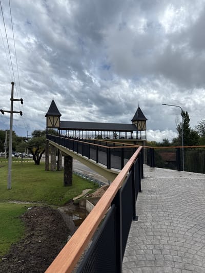 The image depicts a covered pedestrian bridge in Villa General Belgrano, Argentina. The bridge has two prominent towers with pointed roofs. The bridge structure is supported by stone-faced pillars. The bridge has black metal railings and a light-colored wooden handrail. There is a paved walkway on the top right and a grassy area with trees and parked cars visible below the bridge. There are utility poles with power lines in the foreground. The sky is overcast with grey clouds.