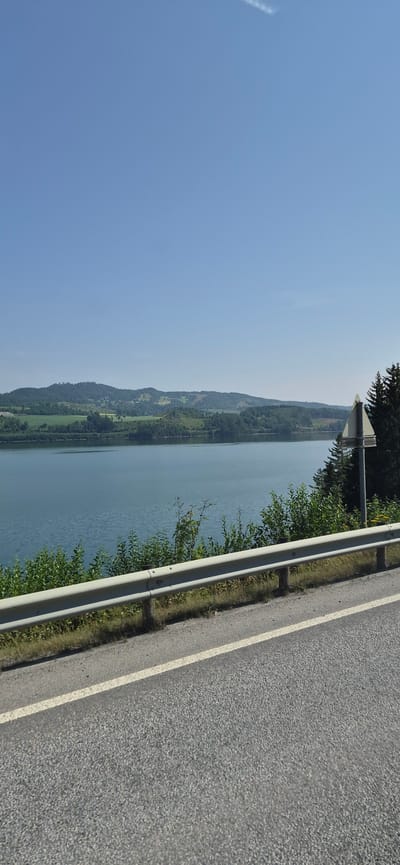 The image shows a scenic view of a body of water with rolling hills in the background. In the foreground, there is a road with a guardrail and some vegetation. The sky is clear and blue. There are no people or specific landmarks visible that would indicate the location in Vingrom, Norway. The scene appears to be a natural landscape with infrastructure elements.