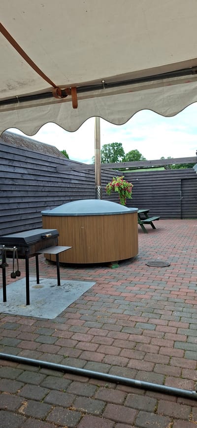 In an outdoor patio area paved with red and gray brick, a round, wooden-paneled hot tub with a gray cover is situated. A metal chimney extends vertically from the center of the hot tub. Adjacent to the hot tub is a black charcoal grill with cooking utensils. In the background, a dark wooden fence encloses the area. To the right of the hot tub, a green picnic table is visible. The scene is partially covered by a white, wavy awning supported by wooden beams. The environment appears to be a private residence or a small commercial establishment in Valma, Estonia.