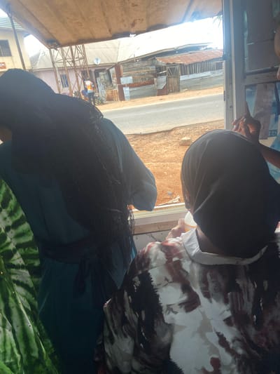 The photo shows two people inside a vehicle or structure in Okengwe - Obehira, Nigeria. One person is wearing a dark head covering. The other is wearing a patterned shirt. Outside the vehicle or structure, there is a street, buildings with corrugated metal roofs, and people in the distance wearing orange and white clothing, possibly construction workers.