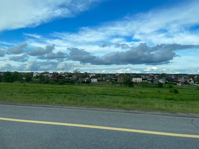 The image depicts a scene in Бавыкино, Russia. It's an upside-down view, showing a blue sky with scattered clouds, a row of houses and trees, a grassy field, and a road with a yellow line. There are no people visible.