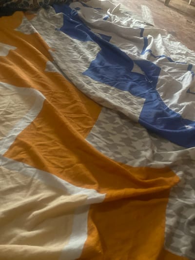 The image shows a close-up, angled view of rumpled bedding with a geometric pattern. The bedding features large blocks of orange and white, intersected by sections of blue and a grey houndstooth pattern. The fabric is creased and folded, suggesting it is a sheet or duvet cover. The lighting is dim, and the overall impression is one of disarray. The location in Kirsinga, Nigeria, is not visually evident from the provided image.