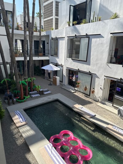 The scene depicts an open-air courtyard of a modern, multi-story building complex located in San José del Cabo, Mexico. The architecture features stark white facades with numerous windows and glass doors, many equipped with black-framed sliding mechanisms resembling barn doors. Tall, slender palm trees are densely interspersed throughout the courtyard and around the perimeter.
In the center of the gravel-covered courtyard is a rectangular swimming pool with dark water, containing a prominent pink, six-ringed inflatable float. Several padded lounge chairs with striped towels are positioned around the pool's concrete edge. To the left of the pool, a single individual is seated on a lounge chair, actively using a laptop computer.
Near the far side of the courtyard, beneath an open white market umbrella, a white reception or display desk is visible. One person is partially obscured behind this desk. To the right of the desk, two individuals stand in an open doorway of one of the buildings; one wears a hat, and the other has lighter hair.
Adjacent to the pool, a cluster of inflatable palm trees in various sizes serves as decorative elements. Potted plants, including cacti and agaves, are visible on balconies and rooftops of the surrounding buildings. The area is brightly lit by natural sunlight, casting clear shadows, indicating daytime.