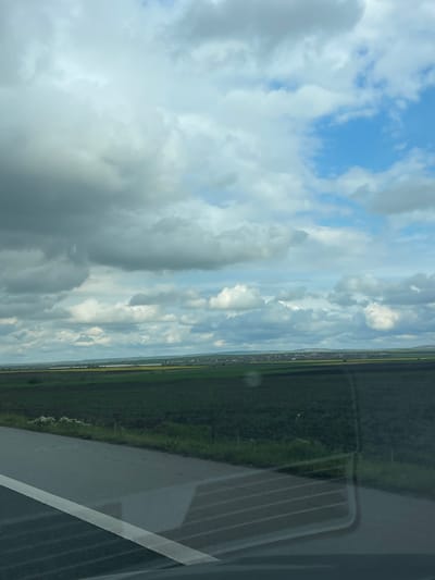 The image, originally rotated 90 degrees clockwise, captures a rural landscape viewed from a vehicle, likely in Vodenichane, Bulgaria, during daytime. The upper two-thirds of the corrected orientation show a vast sky filled with large, fluffy cumulus clouds in shades of white and light grey, interspersed with clear patches of vibrant blue sky.
Below the horizon, the landscape unfolds with extensive green agricultural fields, suggesting a fertile plain. A distinct band of yellow fields, possibly rapeseed, can be seen running horizontally across the mid-distance. Further in the background, a faint line of buildings or a small settlement is discernible against the distant horizon.
The lower right portion of the image is dominated by the reflection of the vehicle's interior on the window. This reflection includes a grey, ribbed structure, possibly an air vent or part of the vehicle's console, and a dark, textured panel. A thick, light-colored line, potentially a road marking or another vehicle component, is also visible within this reflection. The window surface shows some dust or smudges.
The weather appears partially cloudy, with soft, diffused light illuminating the scene. No people are visible, and there is no discernible text in the image. The overall impression is a calm, observational view of a peaceful countryside.