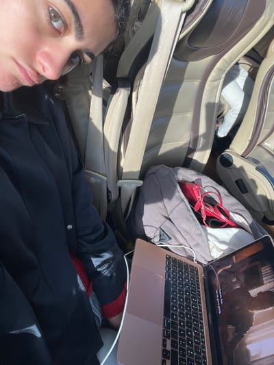 The image captures the interior of a vehicle, located in Castanheira do Ribatejo e Cachoeiras, Portugal, bathed in direct sunlight. A young adult female is visible in the left foreground, secured by a beige three-point seatbelt across her dark-colored jacket and red undergarment. She has dark hair, light brown eyes, and maintains a neutral gaze towards the camera. To her lower right, a rose gold laptop, connected by a white charging cable, is open and displaying video content with visible subtitles. Behind the laptop, a grey bag with an open zipper reveals a red wallet or clutch and red-framed sunglasses. The vehicle's interior features multi-tone beige leather seating with dark brown accents; the lower legs of another passenger in light-colored pants are visible in a seat further back.