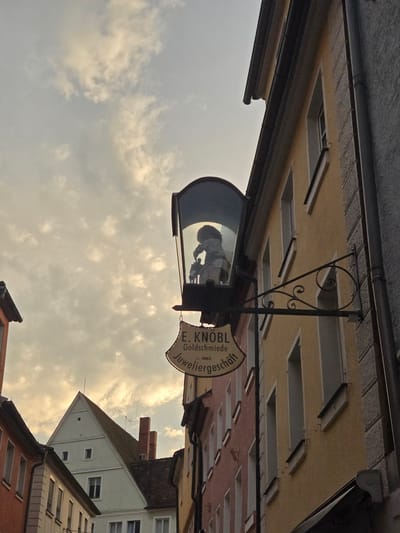 The image depicts a street scene in Ansbach, Germany, focusing on the facade of a building with a hanging sign for "E. KNOBL Goldschmiede Juweliergeschäft". The sign is located above eye level, and beneath it, a decorative display is housed within a glass casing, featuring a silhouette of figures. The building's architecture is visible, with windows and a yellow-toned facade. In the background, other buildings and a triangular-roofed structure are partially visible, under a cloudy sky. The street appears to be narrow, typical of older European towns. The overall lighting suggests it might be late afternoon or early evening.