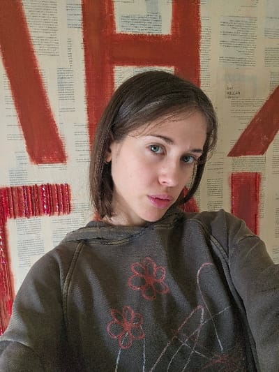 The scene depicts a young adult female located indoors in Doral, United States of America. The subject has fair skin, blue eyes, and shoulder-length dark brown hair parted on the right side. She is wearing a distressed dark grey/brown hoodie adorned with outlines of red flowers and abstract white lines on the front. Her expression is neutral, and she is looking directly into the camera. The background consists of a wall covered with pages of printed text, resembling book pages, overlaid with large, broad strokes of red paint forming partial letters, including elements of an 'H' and potentially an 'A'. Visible text on the wall includes the mirrored phrase 'THE FALLEN ANGEL'. A section on the lower left of the background features multiple horizontal, small, reddish, possibly reflective objects arranged in rows, integrated into the wall art.