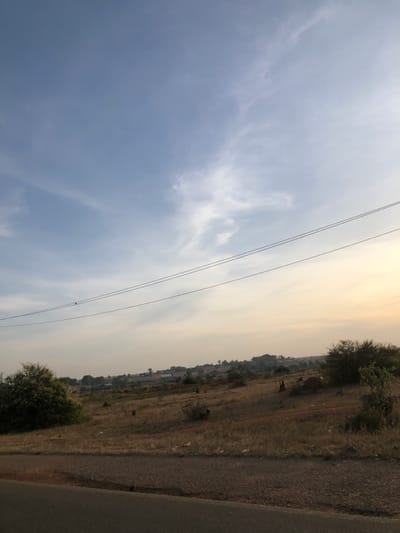 A landscape picture shows a cloudy sky occupying the left half, with power lines stretching across. The right half features a dry, grassy field with scattered trees and vegetation. A road edge runs along the right margin. The horizon is hazy and indistinct, suggesting a dusty atmosphere. The scene evokes a sense of rural expanse under a muted, somewhat overcast sky.