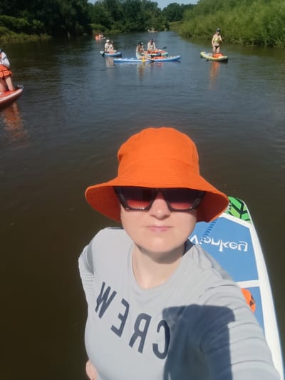 The image shows a woman in an orange bucket hat and sunglasses taking a selfie while on a paddleboard. She is wearing a grey long-sleeved shirt with "CREW" written in large dark letters across the front. The paddleboard she is on is blue and white with green accents and has the word "bouken" visible on it. The woman is in the foreground, and behind her, other people are paddleboarding on a river. The river is surrounded by lush green trees and vegetation on both banks. The sky is blue and clear. The scene appears to be a recreational activity on a warm, sunny day. The location is identified as the city of Berkuty, Russia.