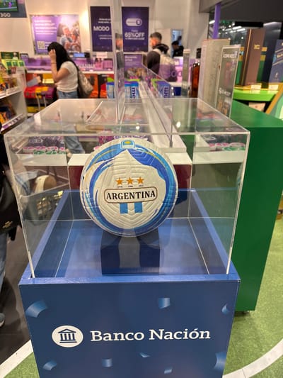 The image shows a display with a soccer ball inside a clear plastic box. The soccer ball is white with blue and gold accents, and features the text "ARGENTINA" prominently. Below the plastic box is a dark blue pedestal with the logo of Banco Nación, an Argentinian bank, and the text "Banco Nación". The background appears to be an indoor retail environment, possibly a store or exhibition, with shelves displaying various products, posters, and other patrons visible. The lighting is bright and the overall scene suggests a promotional or commercial setting. The image does not contain any information that would place it specifically in Ezeiza, Argentina; it only shows the national symbols of Argentina.