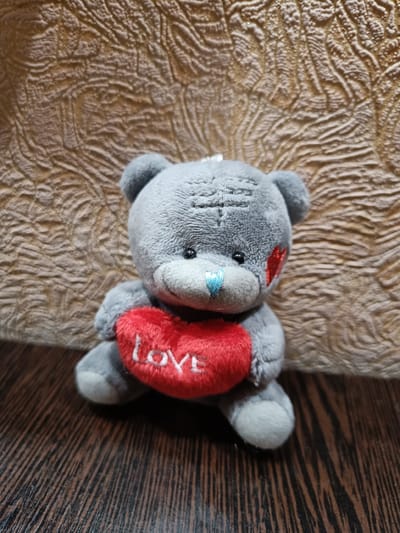A close-up, eye-level shot shows a small, plush gray teddy bear sitting on a dark wooden surface. The bear is facing the viewer and is holding a bright red, plush heart with the word "Love" embroidered in white thread. The bear has a blue stitched nose and red stitched hearts on its cheeks. It has dark eyes and a stitched pattern on its forehead. The background is a textured, light brown surface. The lighting appears to be artificial, casting soft shadows. The overall impression is one of affection and sentimentality. No specific time of day, weather, or strong emotional cues are evident beyond the theme of love suggested by the heart. The provided location context of Vichuga, Russia, is not directly inferable from the visual elements of the image itself.