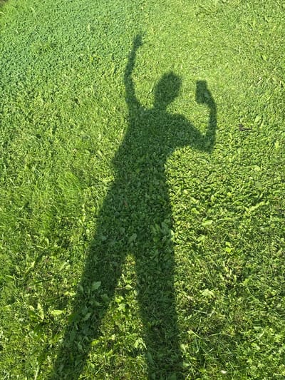 A shadow of a person stands on a grassy area. The shadow is cast from the right side, with the sun illuminating the grass from that direction. The grass is a mixture of various green shades, with some patches appearing lighter and others darker, suggesting varying heights and types of vegetation. Some broad leaves of plants, possibly weeds, are interspersed among the grass blades. The shadow depicts a person with their left arm raised, fist clenched, and their right arm bent, also with a fist clenched. The overall impression is one of dynamic posing, possibly for a photograph. The location in Baryshevo, Russia is noted, but no specific landmarks or urban elements are visible to confirm this.