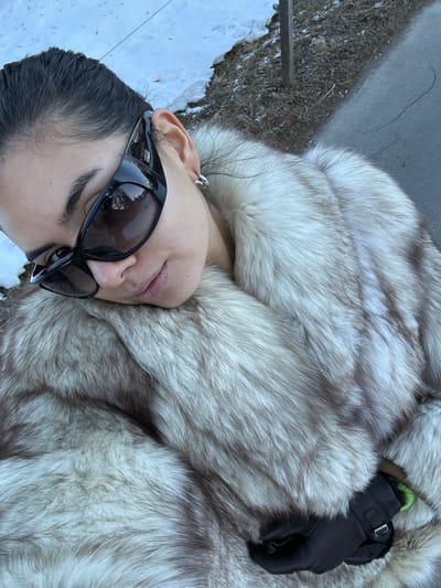 The image displays a partial view of a female subject outdoors in Saanen, Switzerland. The subject's head, with dark hair pulled back, is tilted and rests on a voluminous, multi-toned fur coat primarily composed of light grey and brown fibers. She wears large, dark-tinted wraparound sunglasses that obscure her eyes, and a single silver hoop earring is visible on her right earlobe. Her lips exhibit a subtle upward curve. A black winter glove with a visible lime green interior cuff is present on the subject's left hand, partially concealed by the fur coat in the lower right quadrant of the image. The outdoor environment in the background features patches of snow on brown earth, a dark asphalt path, and a vertical dark-colored pole. Reflections in the sunglasses show indications of trees and sky.