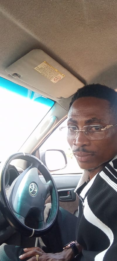 The image captures a close-up portrait of a man seated in the driver's seat of a car, looking directly at the camera. He has dark skin, short, curly black hair, and a neatly trimmed mustache. He is wearing rimless glasses with gold-colored frames that feature an intricate design on the temples. His expression is neutral and direct.
The man is dressed in a black and white garment, possibly a jacket or shirt, with a textured or striped pattern, particularly visible on his right side. On his left hand, a gold-colored ring with a ribbed texture is worn on his ring finger. A black smartwatch with a rose gold or copper-toned case is visible on his right wrist.
The interior of the car is visible around him. A black steering wheel with a silver-toned Toyota logo in the center occupies the lower left portion of the frame. The steering wheel appears to have a cover, and there are signs of wear on its surface. Above the man's head, the car's beige headliner is visible, along with a beige sun visor that is pulled down. A warning label on the sun visor is clearly visible, though mirrored, reading "ADVERTISEMENT" (AVERTISSEMENT) and "WARNING", along with pictograms indicating airbag safety precautions for children. Through the car windows, a very bright, somewhat overexposed outdoor environment is visible, suggesting daylight, with a blue tint at the top of the windshield. A side mirror can be seen through the driver's window. The overall setting is inside a parked car, as the man is posing rather than actively driving.