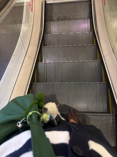 The image shows a first-person perspective of someone standing on an escalator. The escalator's metal steps are visible, as is the side panel of the escalator. The person is wearing sandals and a navy and white striped outfit. They are carrying a green bag with a stuffed animal attached to it. The escalator appears to be in an indoor location. The location is likely in Kelapa Dua, Indonesia.